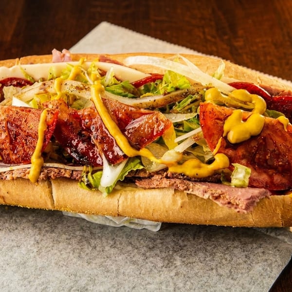 A large, loaded sub sandwich with various meats, cheese, lettuce, onions, and mustard on a wooden table.