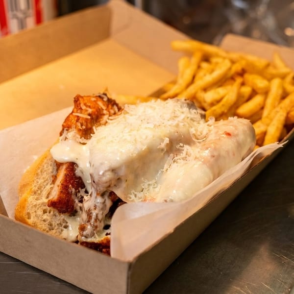 Chicken parmesan sub and french fries in a cardboard takeout container on a metal counter.