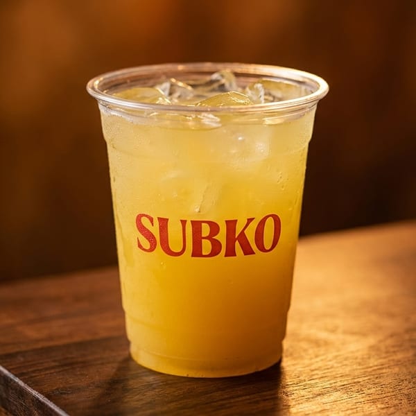 A plastic cup of iced yellow lemonade with the "SUBKO" logo sits on a wooden table.